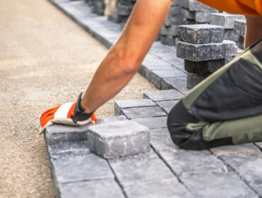 A,Worker,Carefully,Positions,Paving,Stones,On,A,Pathway,,Focusing A worker carefully positions paving stones on a pathway, focusing on alignment and precision. Wearing gloves, the individual kneels on a gravel surface, surrounded by stacks of stone blocks.